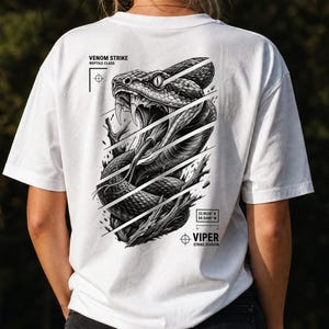 Può includere: Una t-shirt bianca con una grafica in bianco e nero sul retro raffigurante una vipera in attacco con la bocca aperta e la lingua estesa. Il design include il testo VENOM STRIKE, REPTILE CLASS, VIPER STRIKE DIVISION e le coordinate 33.9526 N 84.5499 W.