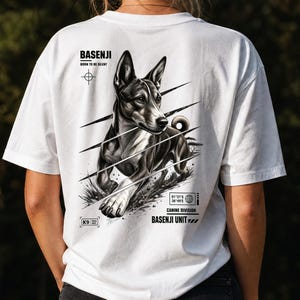 May include: A white t-shirt featuring a black and white graphic on the back of a Basenji dog. The text reads BASENJI BORN TO BE SILENT, K9 FIELD SPEC, 01°21'S 36°49'E, CANINE DIVISION, and BASENJI UNIT. The design includes diagonal lines and a globe icon.