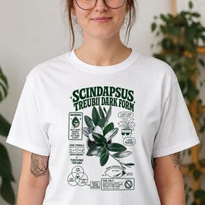 以下が含まれることがあります： Scindapsus Treubii Dark Formの植物が描かれたダークグリーンのグラフィックプリントが施された白い半袖Tシャツ。デザインにはテキスト、植物のイラスト、ドクロの葉やサングラスをかけた植物などのアイコン付きのケア手順が含まれています。