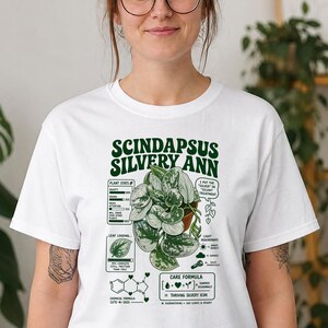 以下が含まれることがあります： Scindapsus Silvery Annの植物の濃い緑色のグラフィックが描かれた白いTシャツ。デザインには植物の統計、ケアの指示、CUTE-N-SASSとラベル付けされた化学式、およびI PUT THE SILVER IN SILENT TREATMENTというテキストが含まれています。