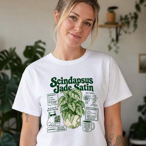 以下が含まれることがあります： 鉢植えのスキンダプサス・ジェイドサテンが描かれた緑色のグラフィック入り白Tシャツ。デザインにはテキスト、サングラスをかけた猫、難易度、成長過程、光の要件、水やりに関するケアの指示が含まれています。