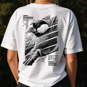 Könnte beinhalten: Ein weißes T-Shirt mit einer schwarz-weißen Grafik auf der Rückseite, die einen Java-Reisfinken auf einem Ast zeigt. Das Design enthält diagonale Streifen, Blätter und den Text Java Sparrow, Small Bird, Big Personality und Finch Division.