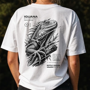 Op de afbeelding: Een wit t-shirt met een zwart-witte afbeelding op de achterkant. Het ontwerp toont een leguaan op een tak met de tekst IGUANA ANCIENT SURVIVORS, REPTILE DIVISION, en een technisch gegevensvak met SPEC: IG-01, LENGTH: 1.8M, WEIGHT: 8.0KG, DIET: HERBIVORE.