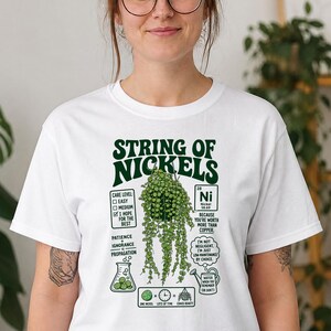 以下が含まれることがあります： String of Nickelsの植物が描かれたダークグリーンのグラフィック入りホワイトTシャツ。デザインにはテキスト、周期表のニッケル元素、ビーカー、じょうろ、植物の世話に関する数式が含まれています。