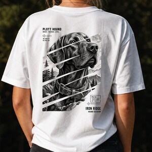 Puede incluir: Una camiseta blanca con un gráfico en blanco y negro de un perro Plott Hound con collar. El diseño incluye líneas diagonales, un fondo de bosque montañoso y el texto Plott Hound, Brave, Focused, Loyal e Iron Ridge Hound Division.