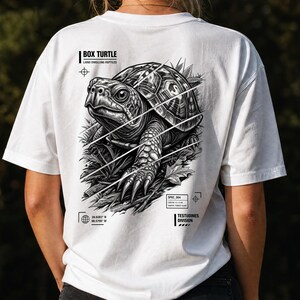 以下が含まれることがあります： 背面にハコガメの白黒グラフィックがプリントされた白いTシャツ。デザインには「BOX TURTLE」、「LAND DWELLING REPTILES」、「SPEC_004」、「TESTUDINES DIVISION」というテキストに加え、座標や技術的なマーキングが含まれています。