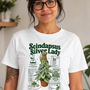 以下が含まれることがあります： 茶色の鉢に入ったスキンダプサス・シルバーレディの植物のグラフィックが描かれた白いTシャツ。デザインにはテキスト、SIシルバーリウムの化学元素ボックス、猫のイラスト、光合成の図、および植物のケア手順が含まれています。