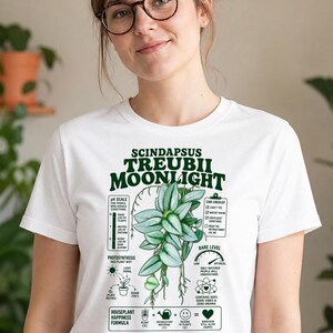 以下が含まれることがあります： Scindapsus Treubii Moonlightの植物の緑色のグラフィックが描かれた白い半袖Tシャツ。デザインにはテキスト、pHスケール、ケアチェックリスト、レア度ゲージ、観葉植物の幸福の公式が含まれています。