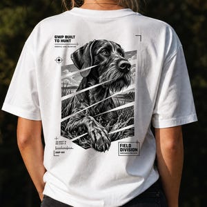 Könnte beinhalten: Ein weißes T-Shirt mit einer schwarz-weißen Grafik eines Deutsch Drahthaar Hundes im Feld. Das Design enthält den Text GWP BUILT TO HUNT, FIELD DIVISION sowie Koordinaten und ist in einem segmentierten, kantigen Rahmen dargestellt.