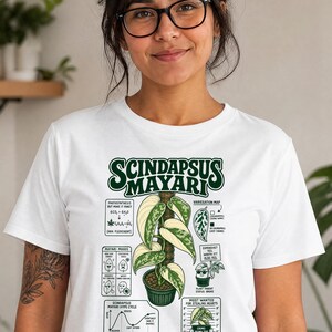 以下が含まれることがあります： 鉢植えのスキンダプサス・マヤリのグラフィックイラストが描かれた白いTシャツ。デザインにはテキスト、光合成の図、斑入りのマップ、ハイプサイクルのグラフ、0から20cmの目盛りが付いた植物のステータスチャートが含まれています。