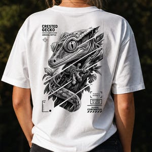 Può includere: Una t-shirt bianca con una grafica in bianco e nero di un geco crestato su un ramo. Il testo riporta CRESTED GECKO, ARBORIAL REPTILE, DIVISION CORONATUS e SIZE RANGE 18-22 CM. Il design include una grafica a righello e delle coordinate.