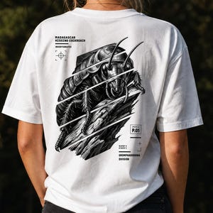 Puede incluir: Una camiseta blanca con una ilustración gráfica en blanco y negro de una cucaracha silbadora de Madagascar sobre un trozo de madera. El diseño incluye etiquetas de texto como Madagascar Hissing Cockroach, Invertebrates, Species ID P.01 y Gromphadorhina Division.