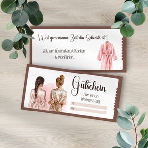 以下が含まれることがあります： 茶色の縁取りが付いた2つのスパデイギフト券。上部のギフト券にはドイツ語のテキストとピンクのローブの水彩イラストが描かれています。下部のギフト券にはドイツ語のテキストと、ローブを着た2人の女性のイラストが描かれています。