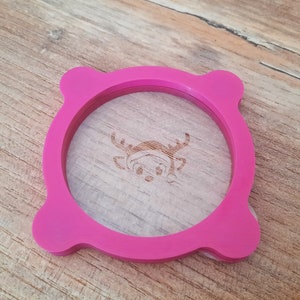 Peut inclure: Un anneau en silicone rose avec un bord surélevé et un motif de renne au centre. L'anneau est conçu pour s'adapter autour d'une tasse ou d'un pot.