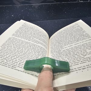 May include: A green plastic page holder for books. The page holder is shaped like a curved rectangle with a hole in the centre for a finger. The page holder is resting on an open book.