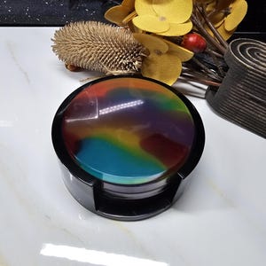 Puede incluir: Un juego de cuatro posavasos coloridos con un soporte negro. Los posavasos tienen un patrón de remolino de azul, verde, amarillo, naranja y rojo.
