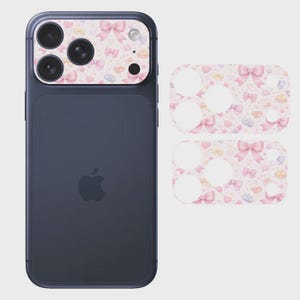 May include: A dark-colored smartphone with a camera lens protector applied to the rear module. The protector features a repeating pattern of light pink bows, small hearts, and tiny blue accents. Two additional identical adhesive protectors are shown.
