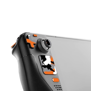 May include: A black handheld gaming device with orange and white accents. The device has a camouflage pattern on the back and a circular button on the top.