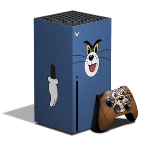 May include: A blue gaming console with a Tom cartoon cat design. The console has a matching controller with a cartoon cat design. The console is rectangular and has a black top with a series of holes.