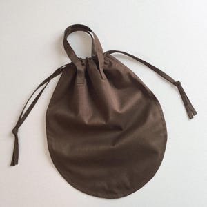 May include: A circular brown fabric pouch featuring a drawstring closure with two long ties and a single top handle. The bag is constructed from a solid-colored brown textile and maintains a rounded shape when cinched at the top with the drawstring.