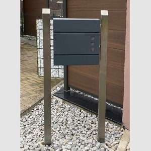 Może przedstawiać: Szara metalowa skrzynka pocztowa z srebrnym metalowym stojakiem. Skrzynka pocztowa ma prostokątny kształt i jest zamontowana na dwóch pionowych słupkach. Skrzynka pocztowa ma szczelinę na pocztę i małe okienko do oglądania zawartości.