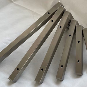 Op de afbeelding: Zes zilveren metalen plankdragers met voorgeboorde gaten. De dragers zijn in een waaier vorm gerangschikt.