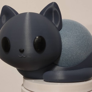 Puede incluir: Una cubierta de altavoz con forma de gato gris oscuro. El gato tiene grandes ojos negros, orejas puntiagudas y una pequeña nariz y boca. El altavoz en sí es una esfera de tela azul claro. El gato está sentado sobre una base blanca.