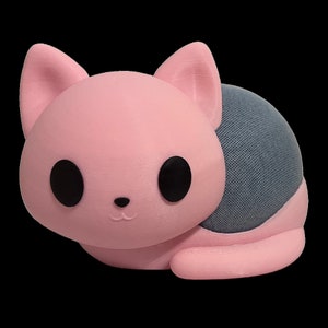 Peut inclure: Un haut-parleur en forme de chat rose avec une grille de haut-parleur en tissu gris à l'arrière. Le haut-parleur a des yeux noirs et un petit nez rose.