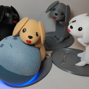 Puede incluir: Cuatro pequeñas figuritas de perros, cada una con un color y diseño diferente, se muestran en bases circulares. Las figuritas se colocan encima de un altavoz inteligente azul con una base de carga blanca.