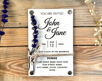 May include: A white invitation card with the text "John & Jane" and event details. The card is decorated with a lavender illustration and secured with metal fasteners. The card is on a wooden surface.