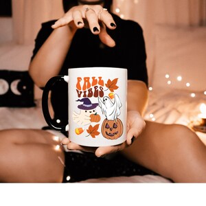 May include: White ceramic mug with a black handle. The mug has a graphic of a ghost wearing a witch hat and holding a pumpkin. The text "Fall Vibes" is printed in a retro font on the mug.