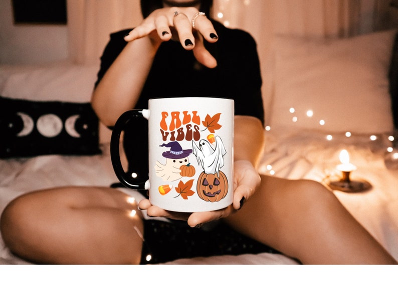 May include: White ceramic mug with a black handle. The mug has a graphic of a ghost wearing a witch hat and holding a pumpkin. The text "Fall Vibes" is printed in a retro font on the mug.