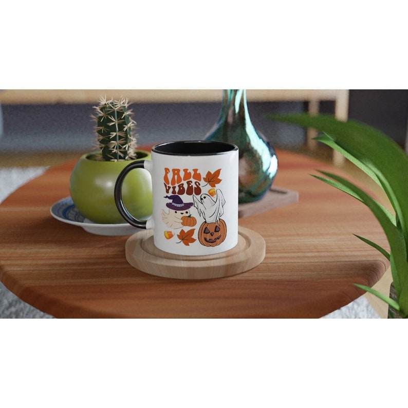 May include: White ceramic mug with a black rim and handle. The mug features a graphic of a ghost and a witch with a pumpkin and the text "Fall Vibes".