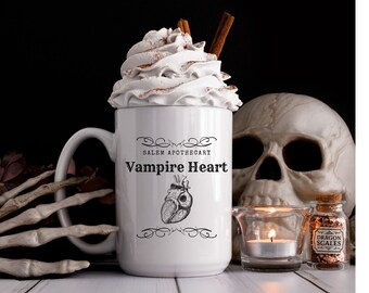 May include: White ceramic mug with a black and white illustration of a heart and the text "Salem Apothecary Vampire Heart". The mug is filled with whipped cream and topped with cinnamon.