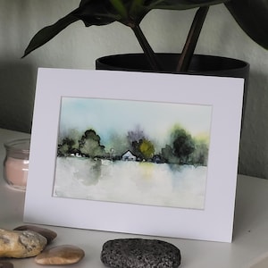 Könnte beinhalten: Aquarell-Landschaftsmalerei mit Bäumen, einem kleinen Haus und Wasser. Das Kunstwerk ist in einem weißen Passepartout gerahmt, mit einer Kerze und Steinen im Vordergrund. Das Gemälde zeigt weiche Blau-, Grün- und Gelbtöne.