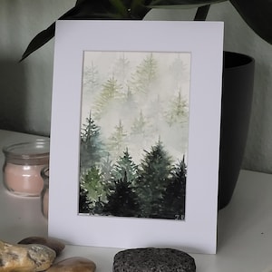 Könnte beinhalten: Aquarellmalerei einer Waldszene, gerahmt in einem weißen Passepartout. Das Kunstwerk zeigt einen nebligen Wald mit immergrünen Bäumen in Grün- und Grautönen. Das Gemälde ist unten rechts signiert. Das Kunstwerk wird auf einer weißen Oberfläche ausgestellt.