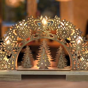 Könnte beinhalten: Weihnachtsdekoration aus Holz mit einem beleuchteten Bogen mit Schneeflocken-Ausschnitten und drei Tannenbäumen. Der warme Schein der Lichter hebt das lasergeschnittene Design hervor. Die Basis besteht aus hellem Holz.