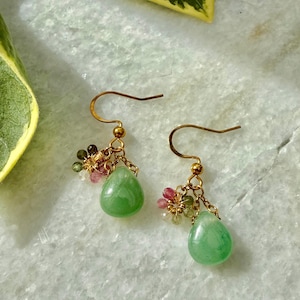 Puede incluir: Un par de pendientes colgantes dorados. Cada pendiente presenta una piedra preciosa verde claro en forma de lágrima, suspendida debajo de un grupo de pequeñas piedras preciosas de colores. Los pendientes tienen un diseño delicado y femenino.