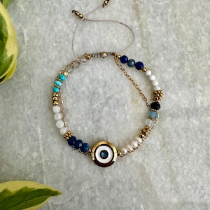 May include: A white and blue beaded bracelet with a gold evil eye charm. The bracelet has a string closure and is made with white, blue, and turquoise beads.