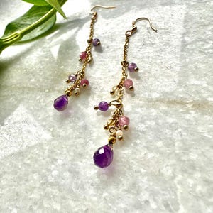 May include: A pair of long, dangling earrings with a gold chain and a variety of purple and pink gemstones. The earrings feature a large, teardrop-shaped amethyst at the bottom.