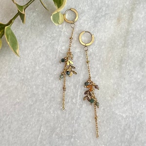 May include: Gold hoop earrings with a long chain and small green, brown, and gold beads and charms hanging from them.
