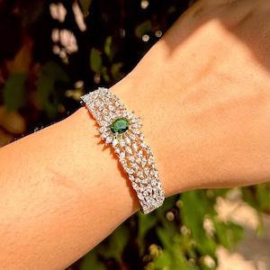 May include: A silver-toned bracelet with a central oval green gemstone, surrounded by smaller white gemstones. The bracelet features a detailed, ornate design with multiple rows of sparkling stones, creating a luxurious look.