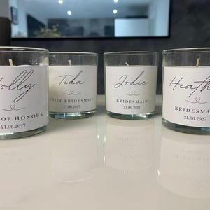 Puede incluir: Cuatro velas de vidrio con etiquetas blancas, cada una con un nombre en escritura elegante: Holly, Tida, Jodie y Heather. Las etiquetas también incluyen títulos como "Dama de honor" y una fecha. Las velas están sobre una superficie blanca reflectante.
