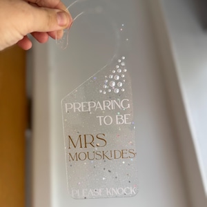 May include: Clear acrylic door hanger with the text "PREPARING TO BE MRS MOUSKIDES PLEASE KNOCK" in gold lettering. The hanger is decorated with glitter and small, clear, round embellishments. The top has a curved handle for hanging.