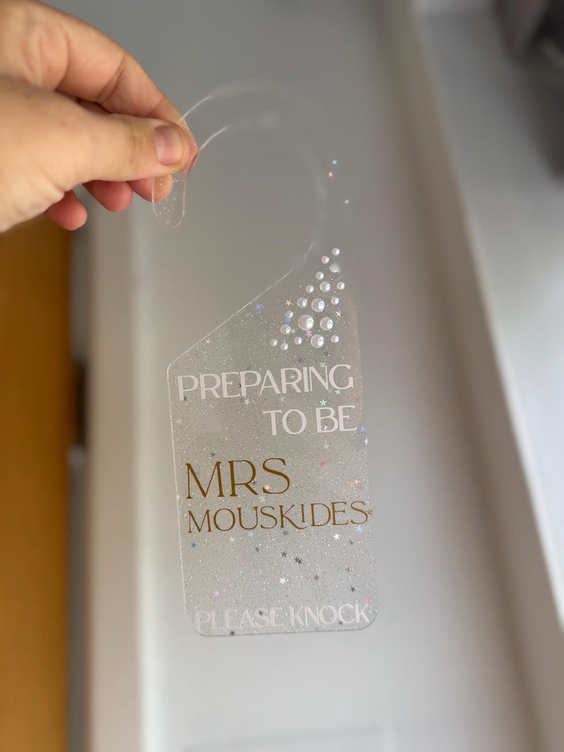 May include: Clear acrylic door hanger with the text "PREPARING TO BE MRS MOUSKIDES PLEASE KNOCK" in gold lettering. The hanger is decorated with glitter and small, clear, round embellishments. The top has a curved handle for hanging.