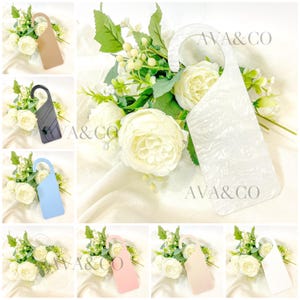 May include: A collection of decorative door hangers in various colors, including beige, black, light blue, white, pink, and cream. Each hanger is displayed with a bouquet of white flowers and the text "AVA & CO".