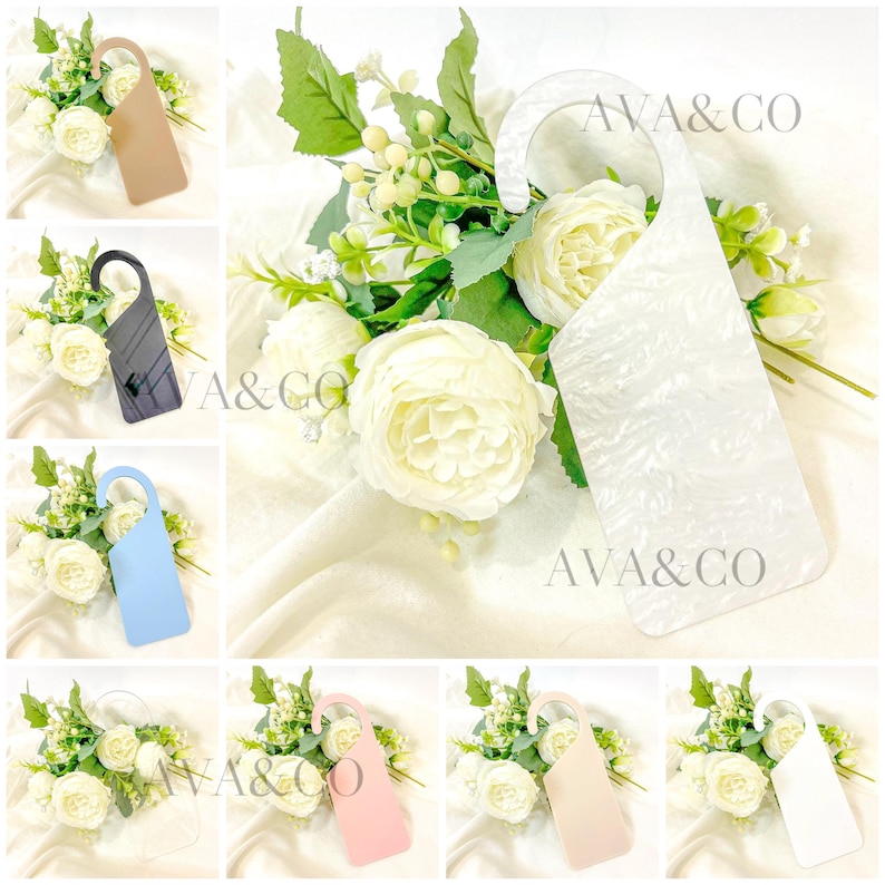 May include: A collection of decorative door hangers in various colors, including beige, black, light blue, white, pink, and cream. Each hanger is displayed with a bouquet of white flowers and the text "AVA & CO".
