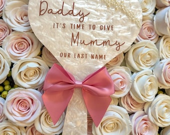 May include: A heart-shaped sign with the words "Daddy, It's Time to Give Mummy Our Last Name" in brown script. The sign has a pink bow and is surrounded by white and pink roses.