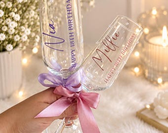 Puede incluir: Dos copas de champán de cristal transparente decoradas con pequeñas perlas blancas. Una copa lleva el nombre Nia y el texto HAPPY 18TH BIRTHDAY en morado. La otra lleva el nombre Millie y el texto HAPPY 18TH BIRTHDAY en oro rosa. Ambas tienen lazos de satén.