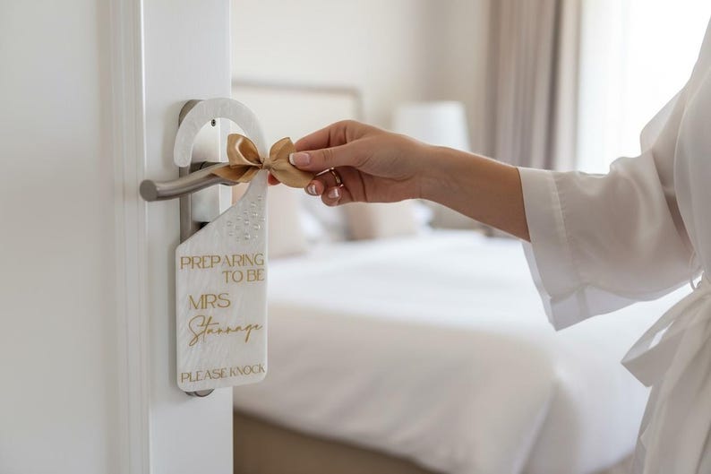 May include: A white door hanger with the text "PREPARING TO BE MRS Stannage PLEASE KNOCK" in gold lettering. A gold ribbon is tied to the top. The door hanger is hanging on a silver door handle.